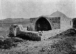 A black-and-white photograph showing a low stone wall enclosing a courtyard in front of a low building with an entry through a pointed arch with a small dome behind