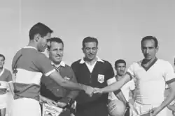 Two football players shaking hands, with two match officials in the background.