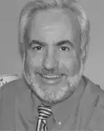 Black and white photo of a middle-aged man with gray hair and beard, wearing a dress shirt and necktie