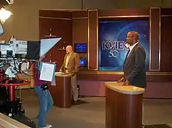 Two men stand at lecterns on a set with KVIE logos in front of a camera