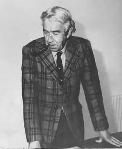 Kenneth Williamson is standing, leaning on a desk. He is wearing a suit and has a pipe in his mouth.