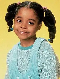 A young African American girl approximately aged five or six smiling at the camera. Dressed in a blue jumper and overalls, her long black hair is worn in a pair of pig tails that protrude from opposite sides of her head.
