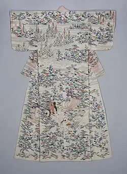 A white kimono with short, rounded sleeves, viewed from the back. The kimono is lined in a coral-coloured fabric, visible through the thin top fabric. The kimono is decorated with an all-over pattern of flowing water, pine trees, plum blossoms, bamboo and decorative rocks, depicted in shades of medium blue, coral, dark purple, grass and emerald green, light pink and gold. Fishing nets are visible at the top of the kimono, and a goshi guruma (flower cart) is seen in the centre of the hem.