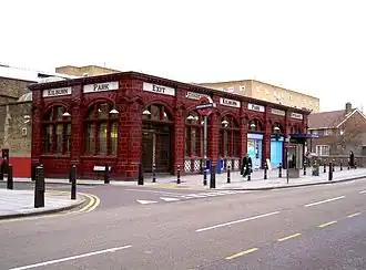 Single storey red glazed terracotta station building with tall arched windows. Glass shaded lamps project from the façade on ornate brackets and the words "Kilburn Park", "Entrance", "Exit" and "Underground" are displayed in black on a white moulded tile band above the window arches.