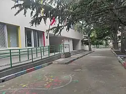 The entrance to a modern, white kindergarten building with a green accessibility ramp. The pavement in front is decorated with colorful painted handprints and chalk drawings.