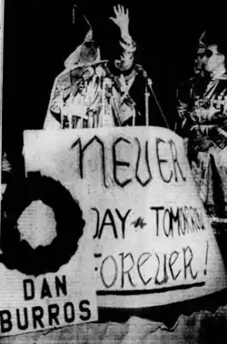 Black and white photo of a man in KKK robes, speaking before a sign. Another sign memorializes Burros