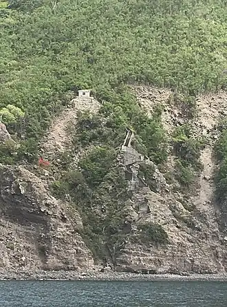 A photo of an island shoreline and cliff, taken from the bay in front of it. Winding up the cliff is a cement staircase leading from the shoreline up to a small white building at the tree-line.