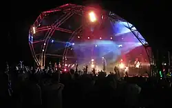 A large stage with a dome extending from its rear and forming a canopy over the front. With blue and red spotlights pointing to a performance. With a packed crowd in attendance.