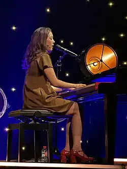 A side-on portrait of Laufey at a concert in 2024. She is illuminated by a spotlight, playing acoustic guitar and singing into a standing mic with her eyes closed.