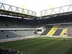 Two-tiered stands with grey seats, below the stands is a pitch with white markings