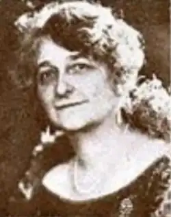 A smiling middle-aged white woman with wavy hair cut in a bob, wearing a dark top with a wide scoop neckline and a strand of pearls