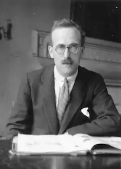 Photograph of a man in middle age, with glasses and a short moustache, reading at a desk.