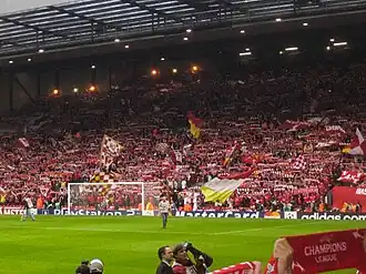 A single-tiered stand that contains thousands of people. Several flags are being waved. In front of the stand is a grass pitch with a goal.
