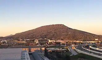 Central tree-covered mountain rising up against a light sky with modern buildings and roads in the foreground