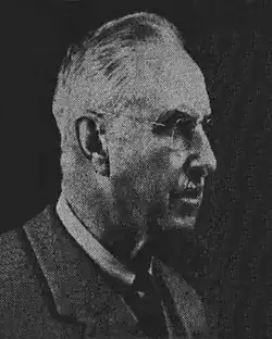 A black-and-white photographic portrait of an older man from the shoulders up, wearing a suit, with a necktie, and eyeglasses