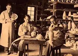 Black and white photograph of the interior of a workshop with three people at work.