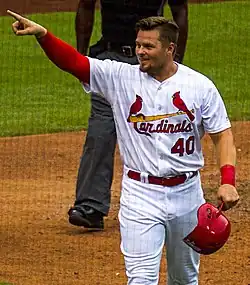 A man in a white baseball uniform with a red right sleeve holding a red helmet in his left hand points with his right hand.