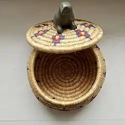 This lidded grass basket was made in 2009 by Annie Novalinga from the community of Sanikiluaq in the Belcher Islands, Nunavut, located in the southeast of Hudson Bay, Canada. The basket is mainly the natural colour of dried grass, and features diamond motifs of blue and red dyed grass on the lid and main basket. The handle is a soapstone carving of an owl.