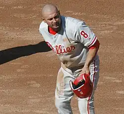 A man in a gray baseball jersey having just removed his red batting helmet at first base. His jersey reads "Phillies" in white and red script and has a number "8" on the left sleeve.
