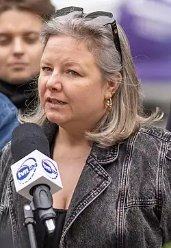 Photo of Broniarczyk speaking into a microphone. She is a white woman with shoulder length salt and pepper hair and a gray denim jacket.