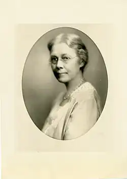 An older woman with silver grey hair pulled back in a bun and round glasses sits in 3/4 profile, looking at the camera. She wears a light-colored blouse and a necklace.