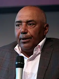 Color photograph of man (Marvin Lewis) wearing a suit and speaking into a microphone.