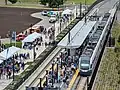 A three-car train is parked at a station with a narrow center platform that is partially covered by a roof. A large crowd of people can be seen on the platform and in an adjacent area with several tents.