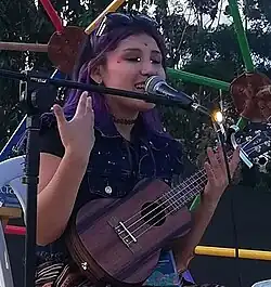 Milena Warthon, seated, with a microphone and ukulele