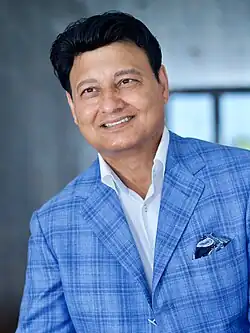 Mohammad Zahoor wearing a light blue checkered suit jacket with a white shirt underneath and a paisley pocket square, smiling, and looking right of the camera