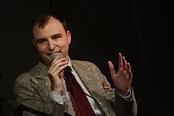 Photo of Jesse Thorn speaking into a microphone. He is caucasian and wearing a coat and tie.