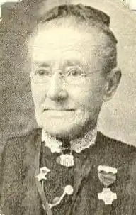 An older white woman wearing glasses; she has medals pinned to her dress.
