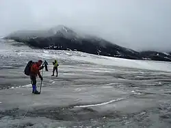 Three climbers descending a glacier on a cloudy day with a rocky mountain peak in the background.
