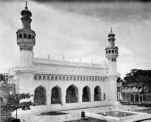 A black and white image of a mosque with five arched entryways, flanked by two minarets on the sides. A fountain is seen in front of the mosque