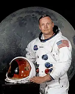 Armstrong posing in his spacesuit, with his helmet on the table in front of him. Behind him is a large photograph of the lunar surface.
