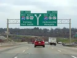 Traffic flows down a four-lane Interstate highway underneath a large sign indicating traffic exits