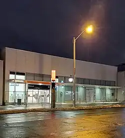 Photo of a light rail station entrance under construction, close to fully completed. It is a low slung building in white with glass panelling and an orange accent.