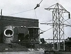 Outside view of brick laboratory building. In the foreground is steel structure, supporting high-voltage lines. The tower is mounted on a base with wheels that ride on rails. Large pieces of equipment are mounted on a steel platform in front of the building.