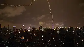 A cityscape with block-like, shadowed towers under a night sky with a bronze glow from a distant illuminated skyline in the background and lightning striking
