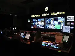 A darkened television control room. Above a bank of monitor displays are a backlit clock and lit signs for CityNews and Citytv. Several people are seated at switchers and computers, controlling the broadcast.