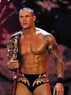 A caucasian male with closely cropped black hair carries a wrestling championship belt over one shoulder. He is wearing short black wrestling tights, with an orange design, and a 'sleeve' tattoo is visible covering one of his arms.