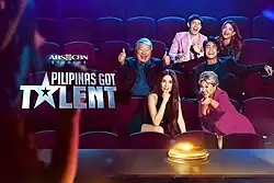 The show's judges and hosts sitting in a movie theatre to the left of the show's logo, facing a table with a glowing golden buzzer