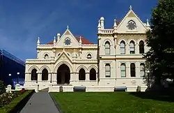 A sunlit grand building is upon a sloping green lawn. It has a big staircase leading to the entrance, several floors and is made of stone in an old fashioned English sort of style. There are flags on flagpoles.