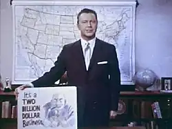 A still shot from the 1965 film Perversion for Profit showing a man in a suit holding a placard. The placard features a cartoonish image of a leering man and the phrase "It's a two billion dollar business"