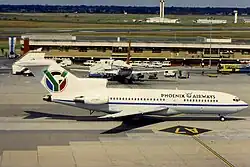A profile view of a white, mid-sized commercial airliner sits on the tarmac at an airport. It has the words Phoenix Airways painted on the side near the front along with a logo on its tail that pays homage to the South African flag.