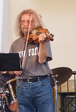 Jiří Kabeš wearing a grey t-shirt that reads "New York" and blue jeans, standing and playing violin in front of a music stand, with drum kit in background