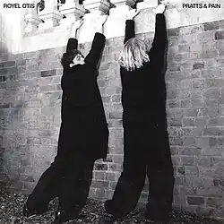 The two band members, wearing baggy all-black clothes, their backs to the camera, lean up against a brick wall and hold onto the balcony above them.