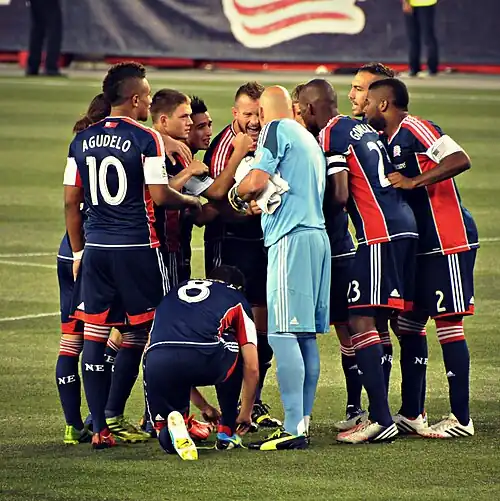 Eleven soccer players in New England Revolution jerseys huddle on the field.