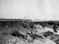 Soldiers march along a sunken dirt road in small groups. Either side of the road debris is strewn and the ground has been churned up from recent artillery attacks