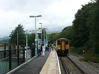 View along country station platform with bilingual exit signage. Passengers wait to board a two carriage train, which is stationary on the single track. Platform, train and track curve slightly right to left. Trees in full leaf stand alongside the track, with mountains in the distance.