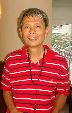 Floro wears a red polo shirt with white and grey stripes. He is wearing a rosary around his neck.
